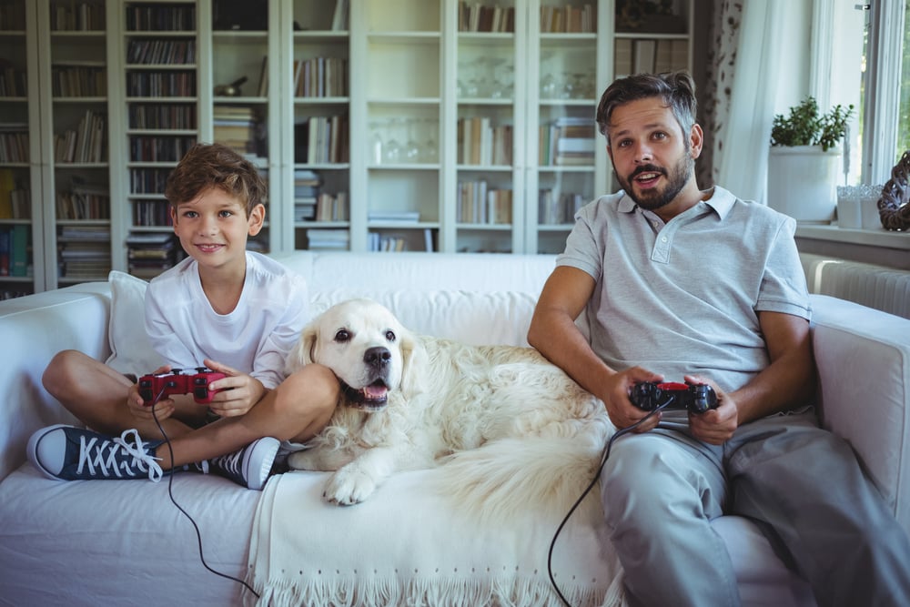 Father and son sitting on sofa with pet dog and playing video games at home